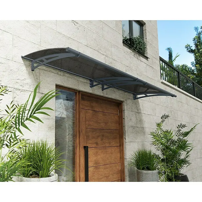 Palram – Canopia Pultvordach Aquila - 302 x 91,5 x 17,5 cm, Anthrazitgrau, Farbe Dach: Solargrau - Nahaufnahme einer grauen Terrassenüberdachung über einer Holztür mit Glaseinsatz, umgeben von üppigen Pflanzen in Töpfen.