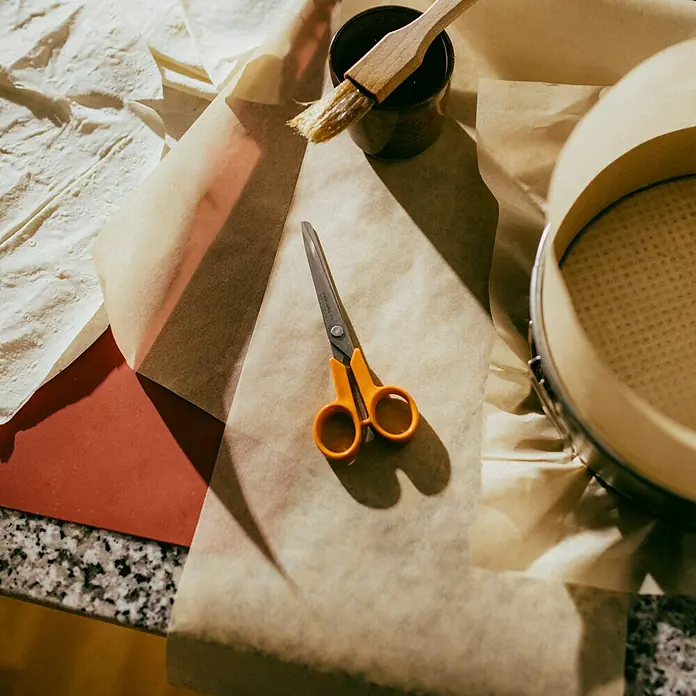 Fiskars Universalschere Classic - Länge: 16 cm, Orange - Küchenutensilien auf Backpapier, darunter eine Schere, ein Pinsel und eine Springform. Vorbereitung zum Backen.