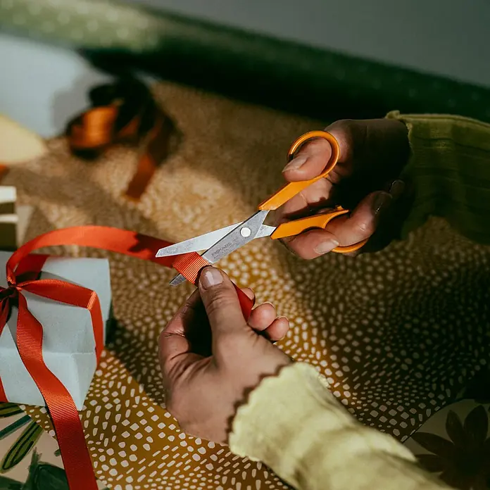 Fiskars Schere Classic - Länge: 13 cm, Orange - Nahaufnahme von Händen, die mit einer orangefarbenen Fiskars-Schere ein rotes Band schneiden, um ein Geschenk zu verpacken.