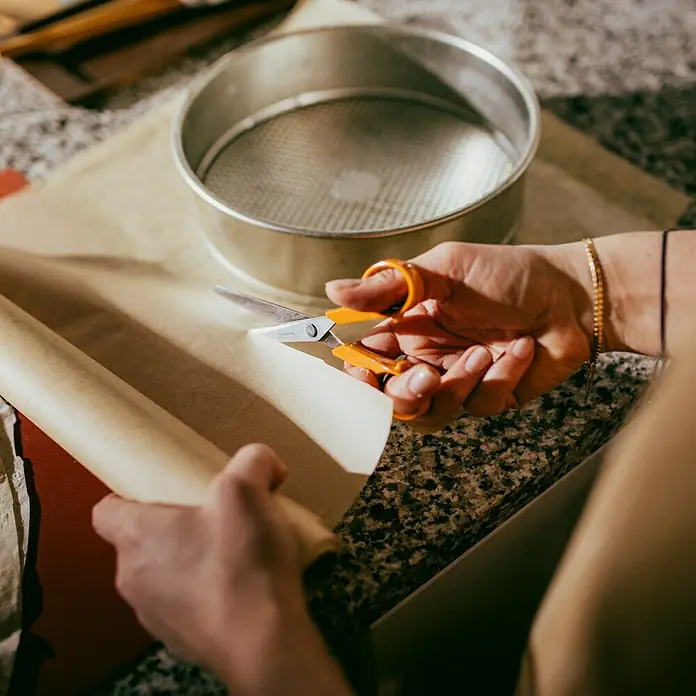 Fiskars Universalschere Classic - Länge: 16 cm, Orange - Ein Küchenszene mit einer Person, die Backpapier mit einer orangefarbenen Schere zuschneidet, neben einem runden Backblech.