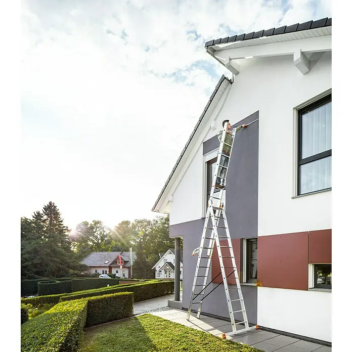 Krause Monto Mehrzweckleiter Tribilo - Arbeitshöhe: 9,3 m, Anzahl Tritte: 36 Stk., Belastbarkeit: 150 kg, Aluminium, Treppengängig - Ein Mann steht auf einer silbernen Leiter und streicht ein Haus mit grauer Farbe. Im Hintergrund sind weitere Häuser und Bäume zu sehen.