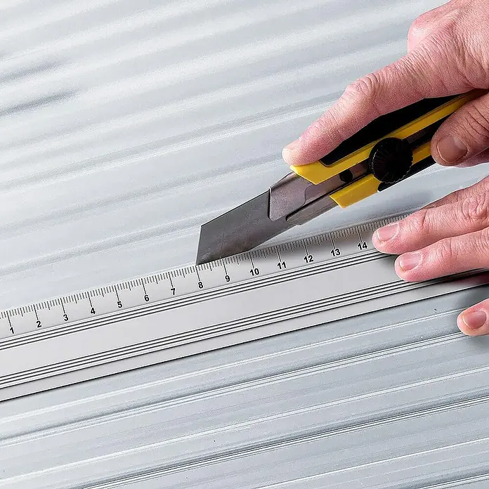 Floordirekt Hohlkammerplatte Doppelstegplatte Polycarbonat 6 mm - 60,5 x 121 cm Transparent -  - Nahaufnahme eines gelben Cuttermessers, das zum Schneiden entlang eines silbernen Lineals auf einer geriffelten Oberfläche verwendet wird.