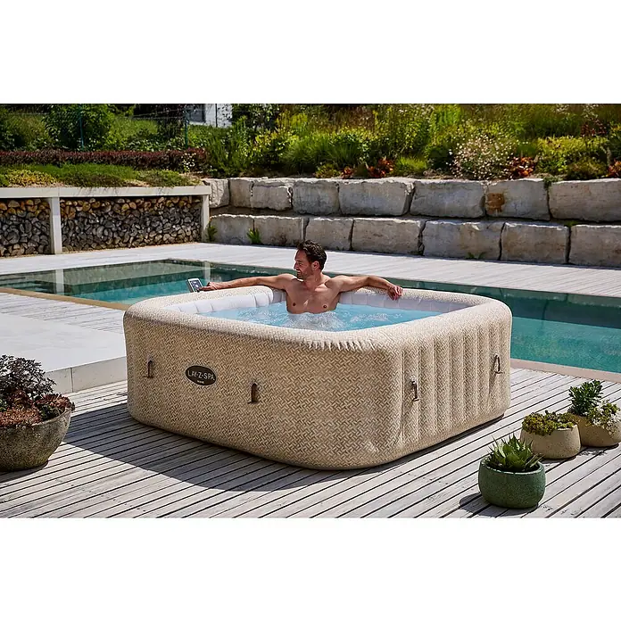 Bestway Lay-Z-Spa Mobil-Whirlpool Rome Air Jet - L x B x H: 201 x 201 x 71 cm, Beige, 1.180 l - Ein Mann entspannt im Lay-Z-Spa Whirlpool im Freien mit Blick auf den Pool und die grüne Landschaft.