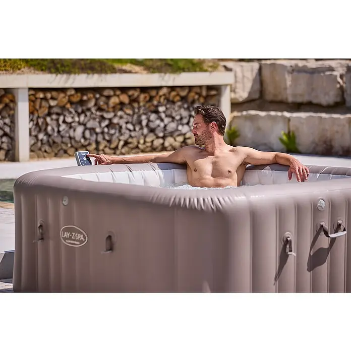 Bestway Lay-Z-Spa Mobil-Whirlpool Copenhagen Hydrojet - L x B x H: 201 x 201 x 80 cm, Grau, 1.050 l - Mann entspannt im Lay-Z-Spa Whirlpool mit Bedienfeld, umgeben von Holz und Steinen. Ideal für erholsame Stunden im Freien.