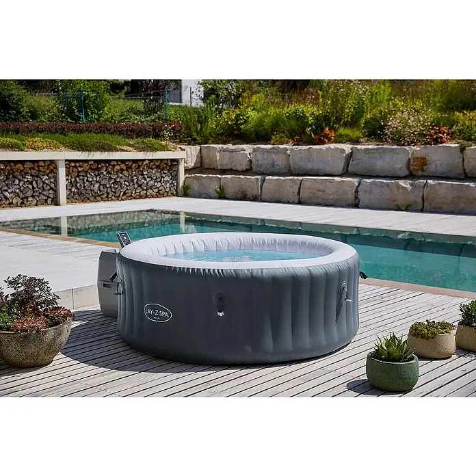 Bestway Lay-Z-Spa Mobil-Whirlpool Miami Air-Jet mit Umrandung - Ø x H: 180 x 66 cm, Anthrazit, 669 l - Ein aufblasbarer Lay-Z-Spa Whirlpool steht auf einer Holzterrasse neben einem Pool und Pflanzen.