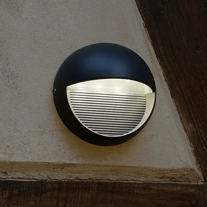 Lutec Aplique exterior LED Radius - 3 W, 17,5 x 17,5 x 10,2 cm, Antracita, Claro, IP54 - Primer plano de una lámpara de pared exterior redonda negra con una cubierta de listones, que emite un brillo cálido y acogedor.
