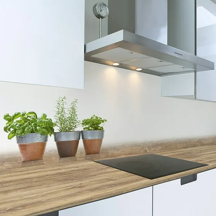 Resopal Küchenrückwand Fixmaß - Cerasum Herbs, 305 x 62 cm, Stärke: 15,4 mm, Holz - Nahaufnahme einer Küchenarbeitsplatte aus Holz mit einem schwarzen Kochfeld und drei Kräutertöpfen im Vordergrund unter einer Dunstabzugshaube.