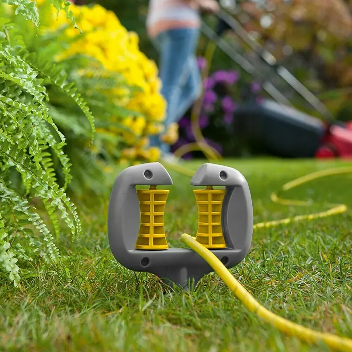 Neptun Schlauch- & Kabelführung - Kunststoff, Erdspieß - Ein grauer Kabelhalter mit gelben Rollen hält ein gelbes Kabel im Garten, während im Hintergrund eine Person den Rasen mäht.