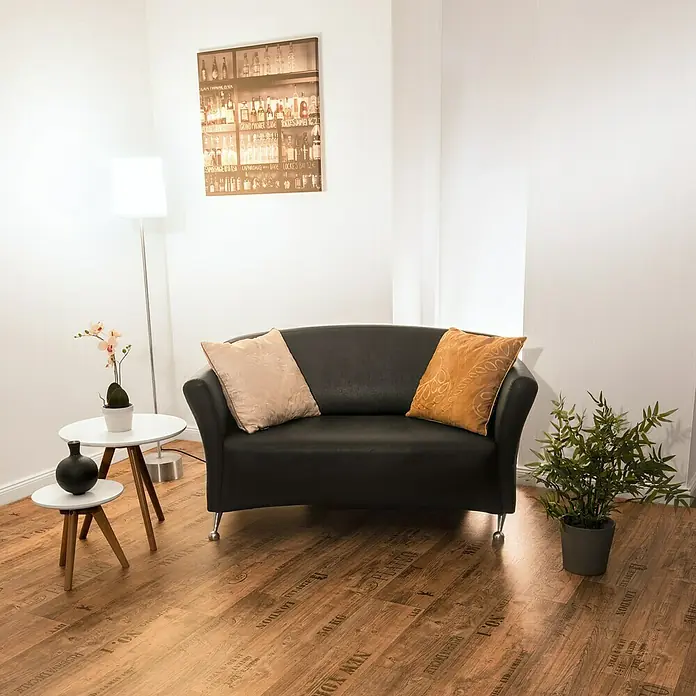CV-Boden Meterware Memphis - 300 cm, Vino, Braun - Schwarzes Sofa mit beigen und gelben Kissen, neben einem Beistelltisch mit einer Vase und einer Orchidee.