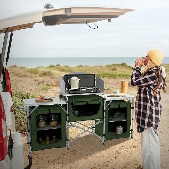 Hauki Campingtisch Camping Küche faltbar mit Tragetasche und Windschutz - L x B x H: 174,5 x 40 x 110,5 cm, Aluminium, 600D Oxford Gewebe, Grau, Khaki - Bild eines grünen Campingkochers mit Stauraum neben einem Auto am Strand, perfekt für Mahlzeiten im Freien.
