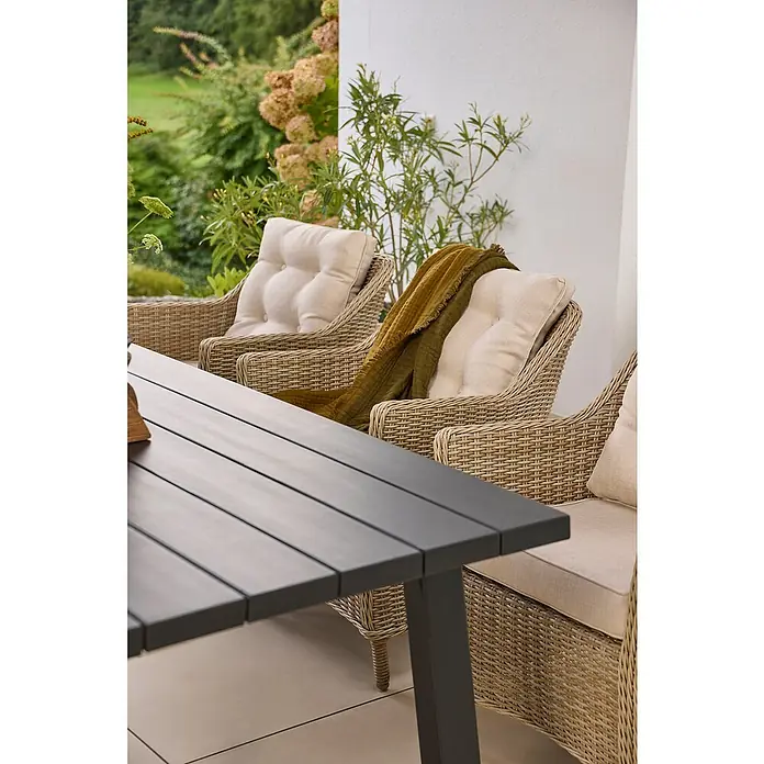 Sunfun Gartenmöbel-Set Melina/Pauline - 5 -tlg., Akazie, Aluminium, Polyrattan, Schwarz/Hellbraun - Ein moderner Gartentisch aus dunklem Holz steht vor Rattansesseln mit beigen Kissen und einer grünen Decke, umgeben von üppigem Grün.