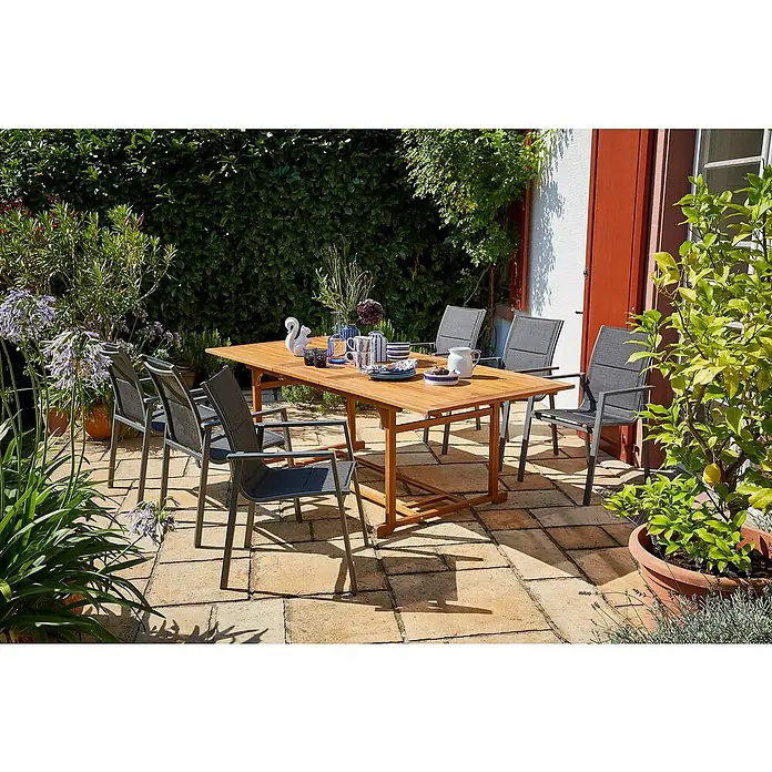 Sunfun Gartenmöbel-Set Diana/Maja - 7 -tlg., Akazie, Aluminium, Textilen, Naturbraun/Grau - Draußen-Szene mit einem Holzesstisch und sechs Stühlen mit grauen Bezügen auf einer Terrasse, umgeben von Grünpflanzen und einem Baum.