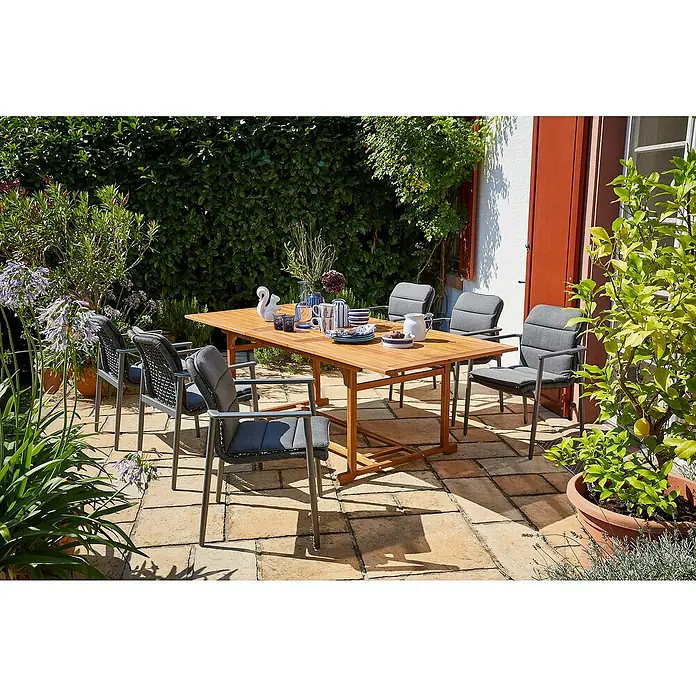 Sunfun Gartenmöbel-Set Diana/Solo Rope - 13 -tlg., Akazie, Aluminium, Olefin, Naturbraun/Night-Grey - Ein Gartentisch- und Stuhlset aus Holz und grauem Polster steht auf einer Terrasse, umgeben von üppigem Grün.