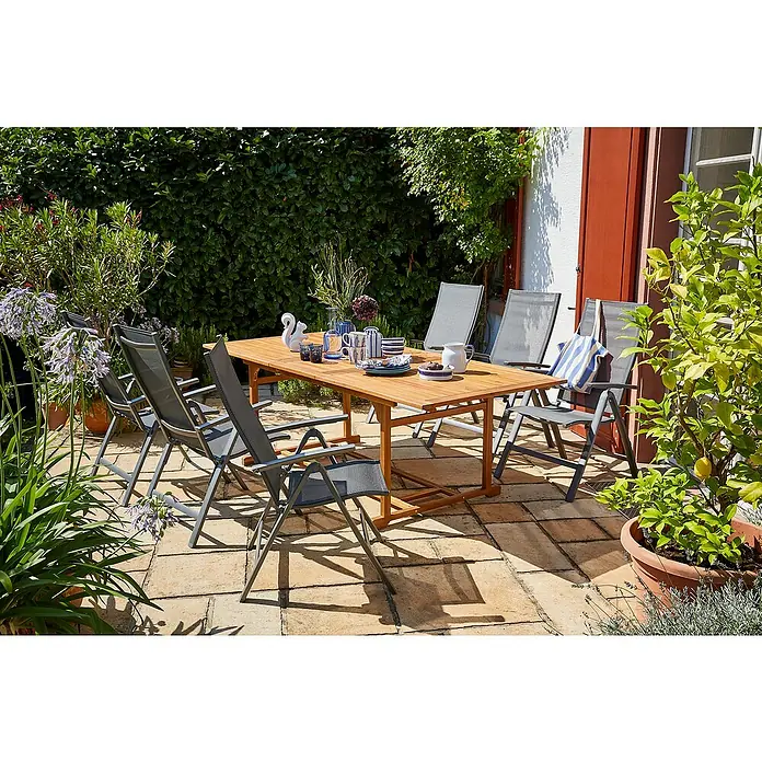 Sunfun Gartenmöbel-Set Diana/Maja Slim - 7 -tlg., Akazie, Aluminium, Textilen, Naturbraun/Anthrazit - Ein Gartentisch aus Holz mit sechs passenden Stühlen auf einer gepflasterten Terrasse, umgeben von üppigem Grün und Pflanzen.
