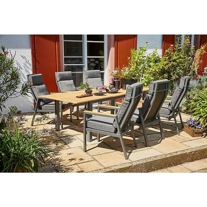 Sunfun Gartenmöbel-Set Carina/Maja Deluxe - 7 -tlg., Aluminium, Stahl, Akazie, Textil, Kunststoff, Night-Grey, Hellbraun - Gartentisch und Stühle auf einer Terrasse mit Pflanzen. Das Set besteht aus einem Holztisch und sechs grauen Stühlen mit Polsterung.