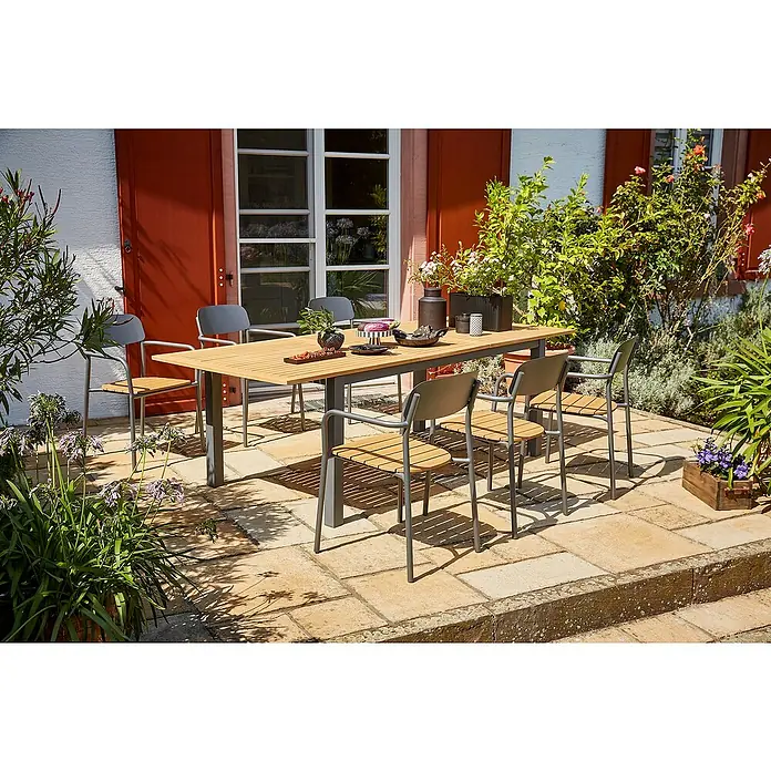 Sunfun Gartenmöbel-Set Carina/Gastro - 7 -tlg., Stahl, Akazie, Polywood, Night-Grey/Hellbraun - Gartenszene mit einem einladenden Esstischset im Freien, umgeben von üppigem Grün. Das Set umfasst einen Holztisch und sechs graue Stühle.
