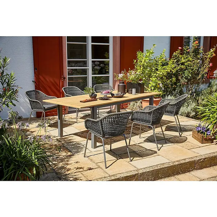 Sunfun Gartenmöbel-Set Carina/Sarah - 7 -tlg., Stahl, Akazie, Olefin, Night Grey/Hellbraun - Gartenset mit einem Tisch und sechs grauen Korbstühlen auf einer Steinterrasse, umgeben von üppigem Grün, ideal für Mahlzeiten im Freien.