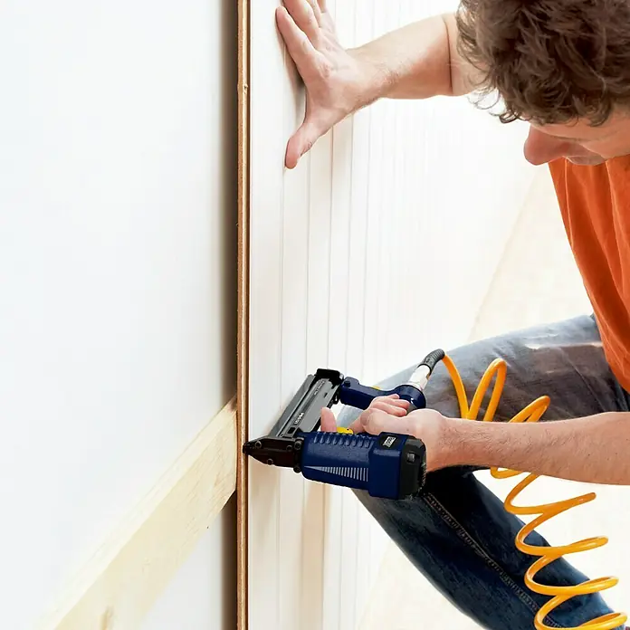 Rapid Kombidrucklufttacker AirTac PBS 121 - Klammertiefe: 12 mm - 30 mm, Geeignet für: Klammertyp 606, Pneumatisch - Ein Mann befestigt eine Holzvertäfelung mit einem blauen Druckluftnagler an einer Wand. Das Werkzeug ist mit einem gelben Schlauch verbunden.