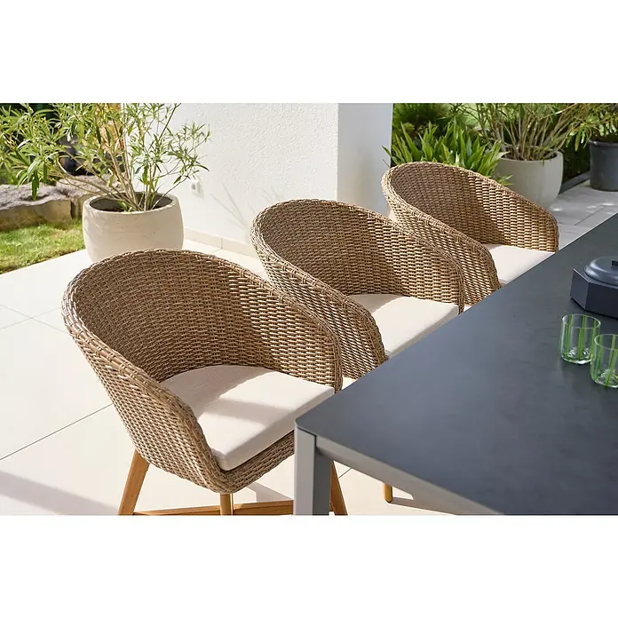 Sunfun Gartenmöbel-Set Maja/Pauline X-Fuß - 7 -tlg., Akazie, Aluminium, Polyrattan, Hellbraun/Night-Grey - Nahaufnahme von drei Korbstühlen mit hellen Polstern und Holzbeinen, die um einen dunklen Tisch auf einer Terrasse stehen.