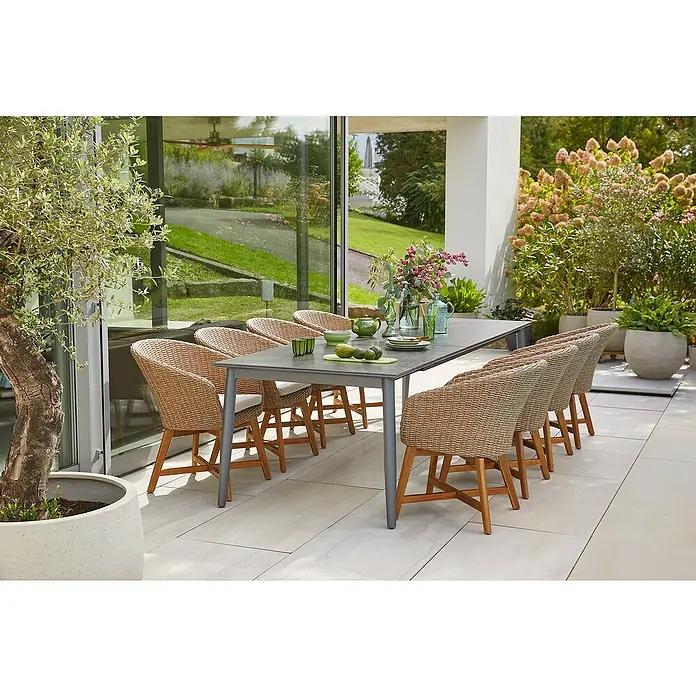Sunfun Pauline Gartensessel - B x T x H: 62 x 70 x 80 cm, Polyrattan, Akazie, Aluminium, Hellbraun - Ein grauer Gartentisch mit Rattanstühlen steht auf einer Terrasse. Dekoriert mit Geschirr, Blumen und Obst. Im Hintergrund üppige Bepflanzung.