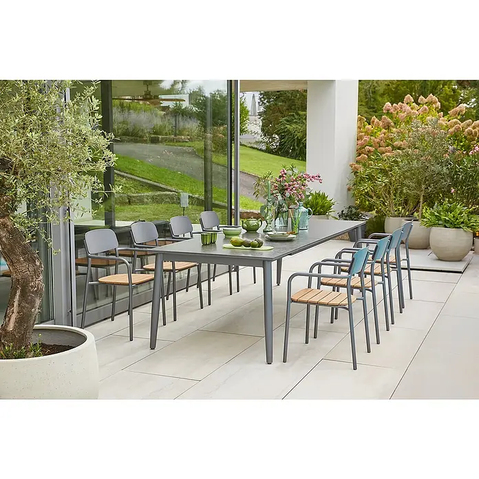 Sunfun Gartenmöbel-Set Solo/Gastro - 9 -tlg., Aluminium, Sintered Stone, Stahl, Polywood, Night-Grey, Tischplatte ausziehbar - Ein grauer Gartentisch mit Stühlen steht auf einer Terrasse mit Pflanzen und Fenstern im Hintergrund. Der Tisch ist mit Geschirr und Obst dekoriert.