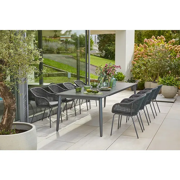 Sunfun Gartenmöbel-Set Solo/Sarah Rope - 9 -tlg., Aluminium, Sintered Stone, Stahl, Olefin, Night-Grey, Tischplatte ausziehbar - Ein moderner Esstisch mit stilvollen Stühlen auf einer Terrasse, umgeben von üppigem Grün. Der Tisch ist mit Geschirr und Dekorationen versehen.