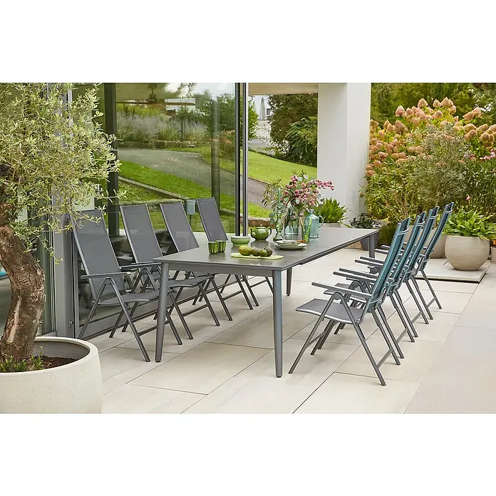 Sunfun Gartenmöbel-Set Solo/Maja Slim - 9 -tlg., Aluminium, Sintered Stone, Textilen, Anthrazit/Night-Grey, Tischplatte ausziehbar - Esstischgarnitur mit einem Tisch und acht Stühlen auf einer Terrasse mit Pflanzen und Garten im Hintergrund.