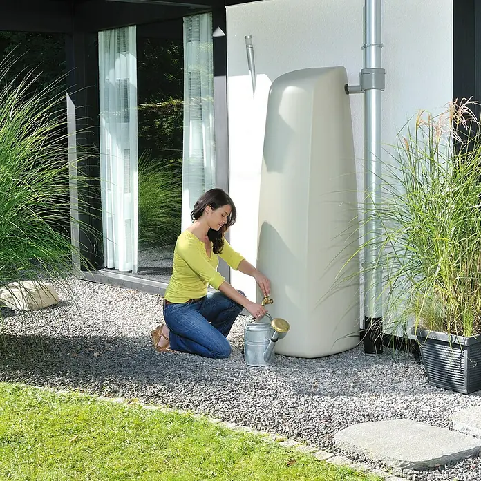 Garantia Classic Line Regenspeicher Elegance - 400 l, Sandbeige - Eine Frau befüllt eine Gießkanne mit einem modernen Regenwassertank vor einem Haus, umweltfreundliche Gartenbewässerung.