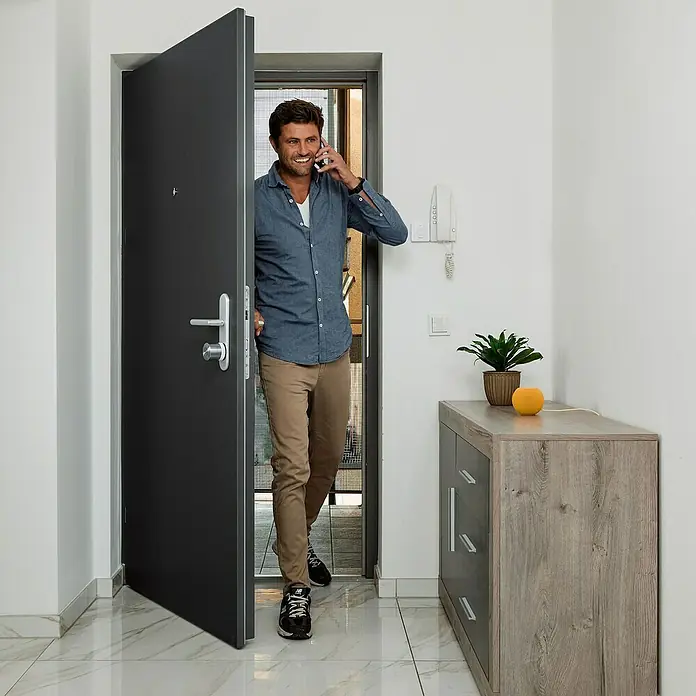 Nuki Elektronisches Türschloss Smart Lock Ultra - Bedienung: Über App, Geeignet für: Türen - Ein Mann betritt ein Haus durch eine moderne, graue Sicherheitstür und telefoniert dabei. Im Hintergrund ist ein Interieur mit einem Schrank sichtbar.