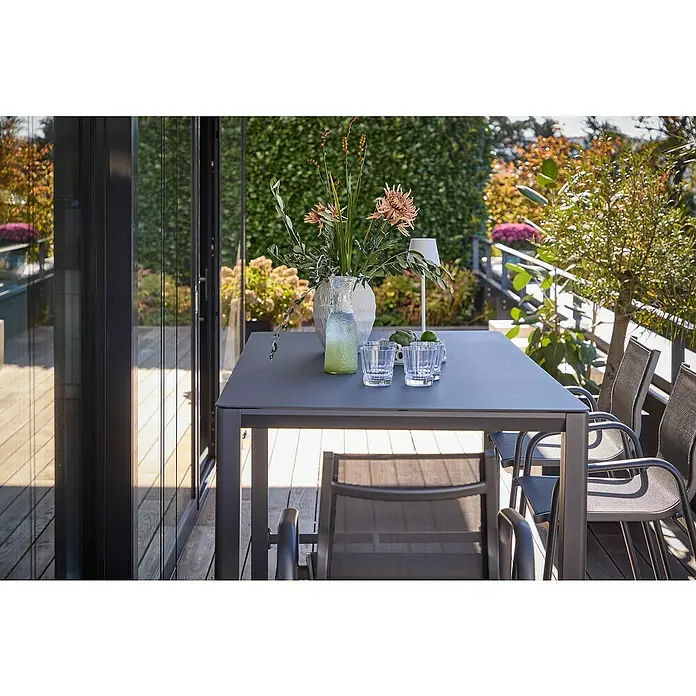 Sunfun Maja Garten-Stehtisch - L x B x H: 160 x 90 x 104 cm, Aluminium, Night-Grey - Ein grauer Esstisch mit passenden Stühlen steht auf einer Terrasse. Auf dem Tisch befinden sich Gläser, Blumen und Dekorationen.