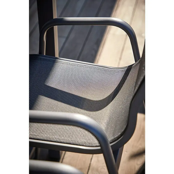 Sunfun Gastro Garten-Barhocker - Stahl, Textilen, Stapelbar, Night-Grey, B x T x H: 53,5 x 74 x 110 cm - Nahaufnahme eines grauen Stuhls mit Netzgewebe-Sitz und Armlehnen, der auf einer Holzterrasse steht. Die Sonne wirft einen Schatten auf den Stuhl.