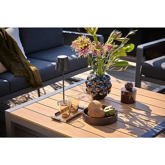 Sunfun Maja Loungetisch Holzoptik - L x B x H: 100 x 100 x 42 cm, Aluminium, Night-Grey - Ein stillvoller Couchtisch mit einer eleganten Lampe, Gläsern, einer Blumenvase und einer dekorativen Schale mit Früchten im Freien.
