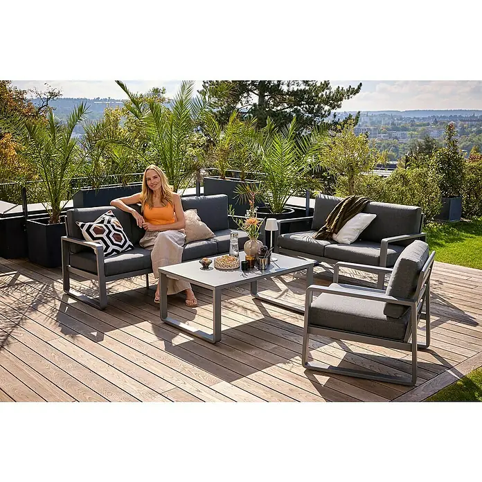 Sunfun Maja Loungesofa 3-Sitzer - B x T x H: 203 x 76 x 74,5 cm, Aluminium, Olefin, Night-Grey - Eine Frau entspannt auf einer grauen Gartenlounge mit Tisch und Stuhl auf einer Holzterrasse, umgeben von Pflanzen.