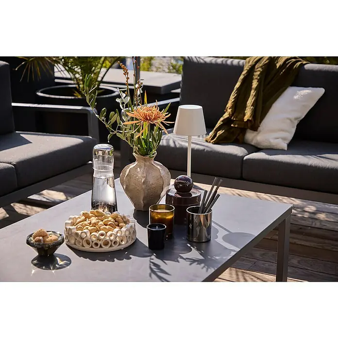Sunfun Maja Loungetisch - L x B x H: 110 x 73 x 46 cm, Aluminium, Sicherheitsglas, Night-Grey - Gartentisch mit Snacks, Getränken und Dekorationen. Der Tisch ist Teil einer Lounge-Ecke mit einem dunklen Sofa und Kissen.