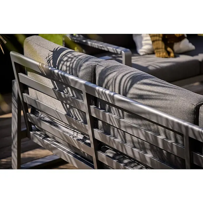 Sunfun Maja Loungesofa 3-Sitzer - B x T x H: 203 x 76 x 74,5 cm, Aluminium, Olefin, Night-Grey - Nahaufnahme eines modernen Sofas mit grauem Stoffbezug und einem eleganten Metallrahmen unter dem Schatten einer Pflanze.