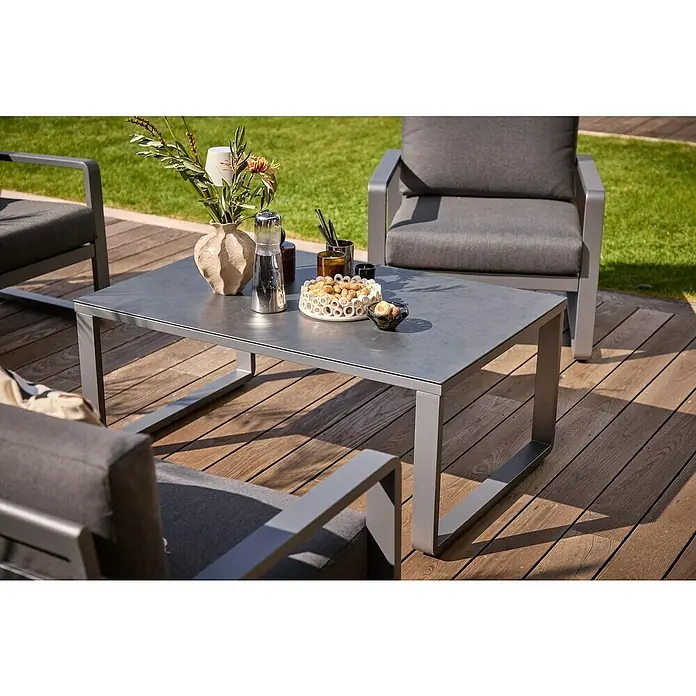Sunfun Maja Loungetisch - L x B x H: 110 x 73 x 46 cm, Aluminium, Sicherheitsglas, Night-Grey - Ein grauer Couchtisch mit Snacks und Dekorationen auf einer hölzernen Terrasse, umgeben von Sesseln mit grauen Kissen für den Außenbereich.