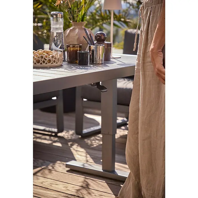 Sunfun Maja Garten-Loungetisch - L x B x H: 138,5 x 70 x 72 cm, Stahl, Night-Grey - Ein verstellbarer Esstisch im Freien, gedeckt mit Geschirr und Dekorationen, steht auf einer Holzterrasse. Die Tischhöhe ist durch einen Hebel anpassbar.