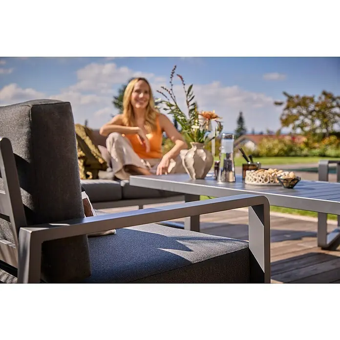 Sunfun Maja Loungesofa 2-Sitzer - B x T x H: 139,5 x 76 x 74,5 cm, Aluminium, Olefin, Night-Grey - Nahaufnahme eines grauen Gartenstuhls mit passendem Tisch, einer Frau und Dekorationen im Hintergrund. Komfortable und stilvolle Outdoor-Möbel.
