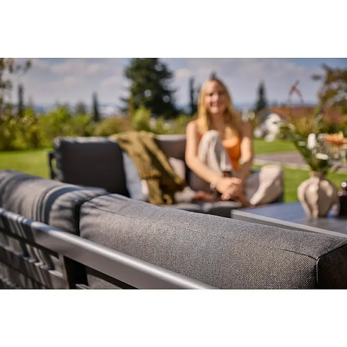 Sunfun Maja Loungesofa 2-Sitzer - B x T x H: 139,5 x 76 x 74,5 cm, Aluminium, Olefin, Night-Grey - Nahaufnahme einer grauen Outdoor-Sofagarnitur mit einer Frau im Hintergrund. Das Sofa lädt zum Entspannen im Freien ein.