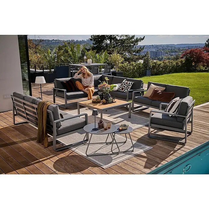 Outdoor-Teppich Newberry - Grau, 230 x 160 cm, 100 % Polypropylen - Terrassenmöbel-Set mit grauen Polstern und Aluminiumrahmen, bestehend aus Sofas, Sesseln und Tisch auf einer Holzterrasse mit Grünblick.