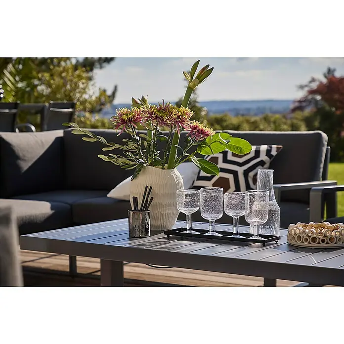 Sunfun Maja Garten-Loungetisch - L x B x H: 138,5 x 70 x 72 cm, Stahl, Night-Grey - Nahaufnahme eines grauen Gartentisches mit Blumen, Gläsern und Snacks vor einem Sofa, der eine entspannte und stilvolle Outdoor-Szene zeigt.