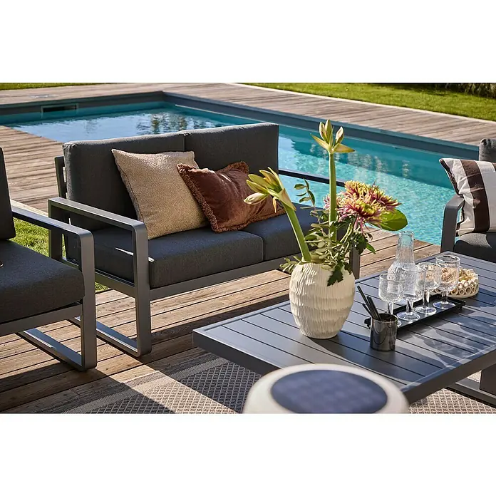 Sunfun Maja Gartentisch - L x B x H: 138,5 x 70 x 72 cm, Stahl, Night-Grey - Nahaufnahme eines grauen Outdoor-Sofas mit Kissen und einem Couchtisch mit Blumen und Getränken neben einem Pool.