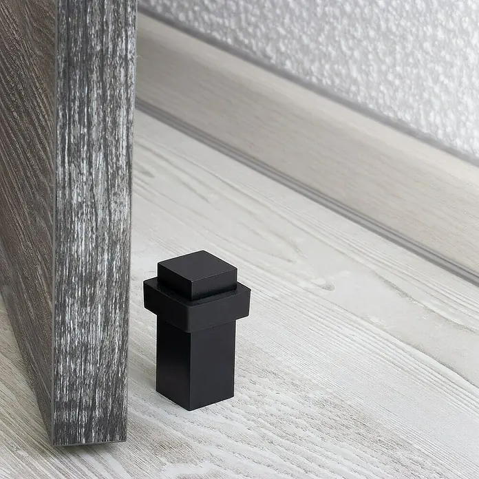 Walteco Bodentürstopper - L x B x H: 20 x 20 x 48 mm, Metall - Nahaufnahme eines schwarzen, würfelförmigen Türstoppers aus Metall, der neben einer Tür auf einem Holzboden steht.
