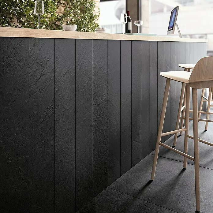 Feinsteinzeugfliese Lavagna Nera - 60 x 120 cm, Schwarz, Matt - Detailaufnahme einer Theke mit stilvollen, schwarzen Steinpaneelen und hellen Holzelementen, ergänzt durch moderne Barhocker für ein elegantes Ambiente.