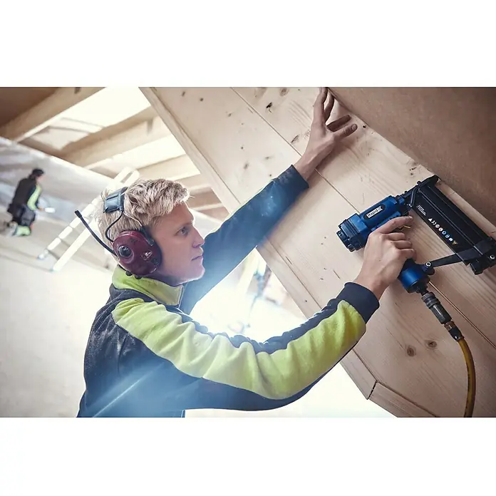 Rapid Nagel Typ 8 - Länge: 30 mm, 5.000 Stk., Verzinkt - Ein Handwerker befestigt Holz mit einem blauen Druckluftnagler, während er Kopfhörer trägt, in einer unfertigen Dachkonstruktion.
