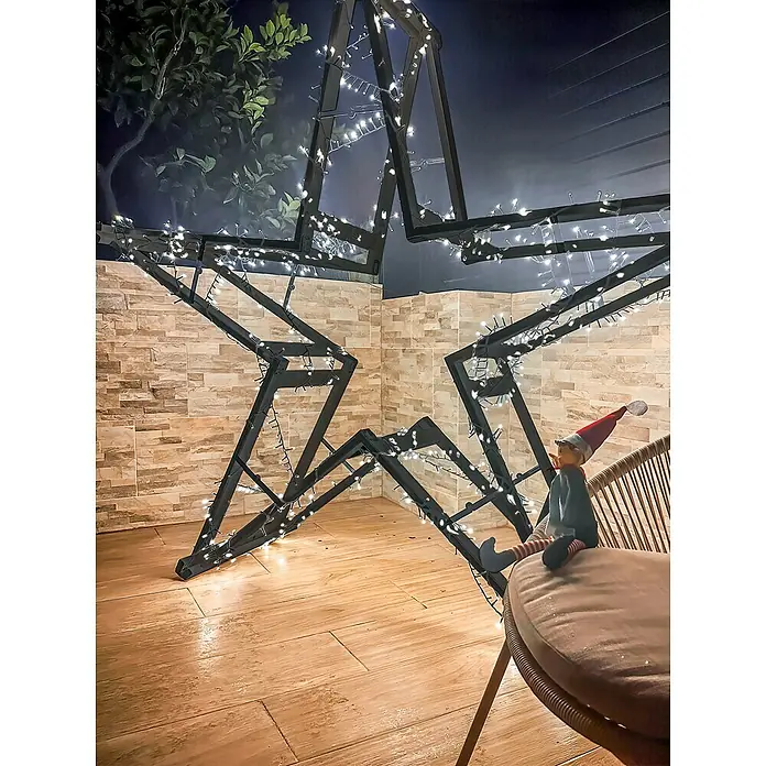 Astigarraga Holzklappbock Standard - Tragkraft: 400 kg, Höhe: 74 cm, Kiefer - Dekorative Lichterkette in Sternform, die eine gemütliche Atmosphäre schafft, während ein festlicher Elf auf einem Stuhl sitzt.