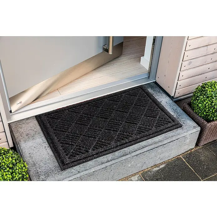 Fußmatte Nub Star - Anthrazit, 63 x 43 cm, 90 % Polyester, 10 % Polypropylen - Eine schwarze Fußmatte mit einem geometrischen Muster liegt vor einer offenen Tür auf einer betonierten Stufe.