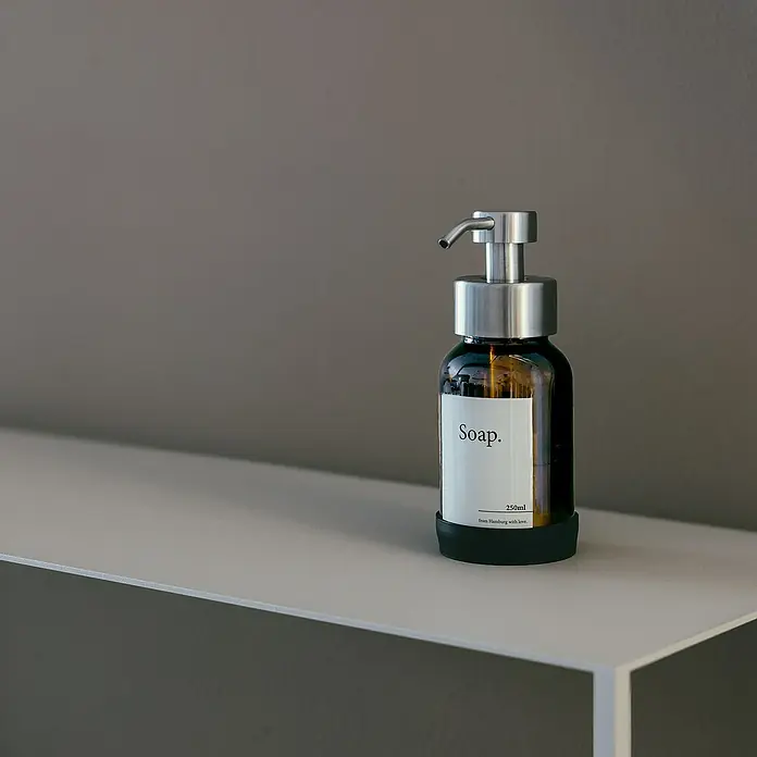 Designfabrik Hamburg Seifenspender - Edelstahl, Silber/Braunglas - Braune Seifenflasche mit silberner Pumpe und weißem Etikett "Soap." auf einem weißen Regal vor grauem Hintergrund.