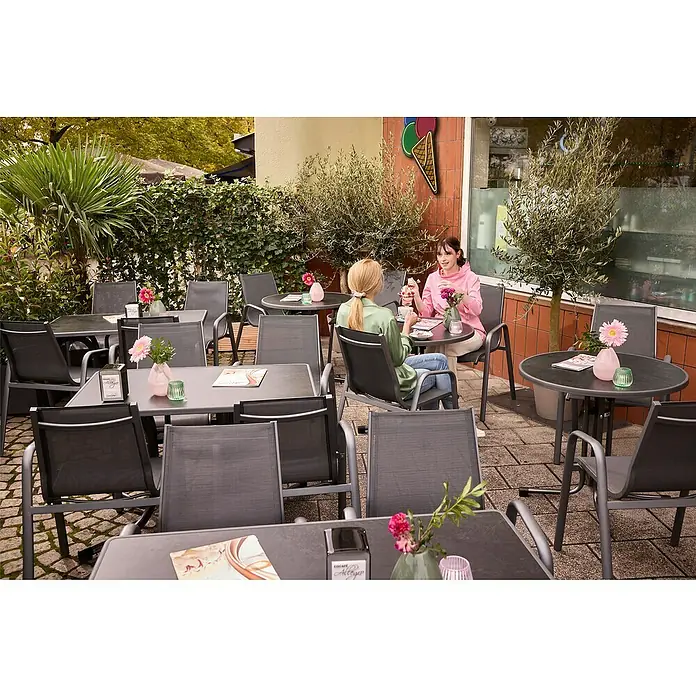 MFG Gartentisch Boulevard - L x B: 110 x 70 cm, Stahl, Mecalit, Schiefer, Klappbar - Straßencafé mit schwarzen Tischen und Stühlen, zwei Frauen sitzen an einem Tisch. Dekoration mit Blumen und Eis-Logo an der Wand.