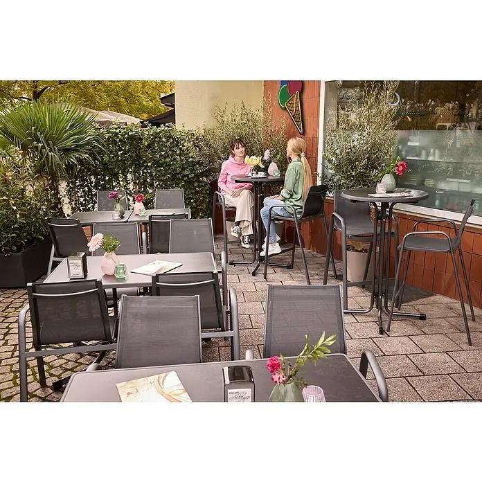 MFG Garten-Stehtisch rund - Ø x H: 70 x 110 cm, Stahl, Mecalit, Rund, Schiefer - Einladende Terrasse mit Tischen und Stühlen vor einem Café, dekoriert mit Blumen. Eine entspannte Atmosphäre für Gäste zum Verweilen.
